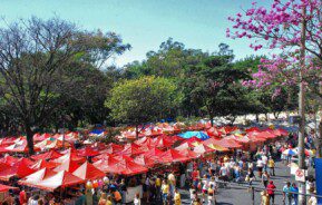 Foto que ilustra matéria sobre a Feira Hippie BH mostra dezenas de barracas, uma ao lado da outra, a maioria delas com coberturas vermelhas, com uma área arborizada de um lado um pedaço do asfalto da avenida Afonso Pena do outro, em um dia ensolarado e com muitos visitantes na feira.