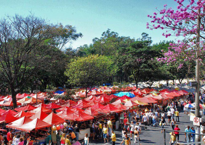 Foto que ilustra matéria sobre a Feira Hippie BH mostra dezenas de barracas, uma ao lado da outra, a maioria delas com coberturas vermelhas, com uma área arborizada de um lado um pedaço do asfalto da avenida Afonso Pena do outro, em um dia ensolarado e com muitos visitantes na feira.