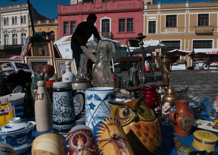 Foto com diferentes artesanatos, principalmente vasos em cores e tamanhos diversos. Ao fundo, há quadros de pintura e, mais adiante, construções históricas.