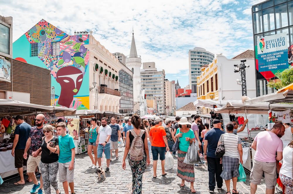 Foto que ilustra matéria sobre feiras em Curitiba mostra muitas pessoas andando em uma rua de paralelepípedos cercada de sobrados em meio a barracas de feiras no Largo da Ordem, em Curitiba.