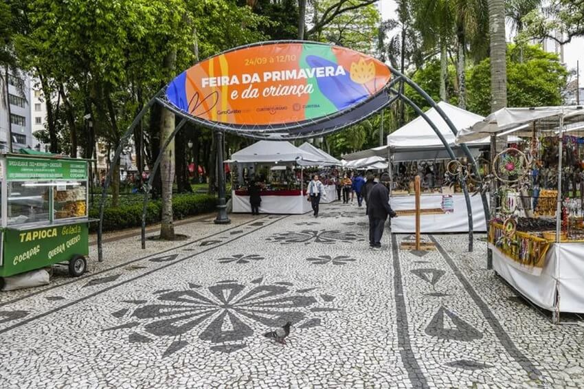 Foto que ilustra matéria sobre feiras em Curitiba mostra barracas montadas em uma praça arborizada, com um portal de entrada para a Feira da Primavera e da Criança