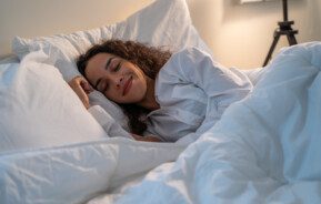 Mulher dormindo em uma cama com roupa de cama toda branca.