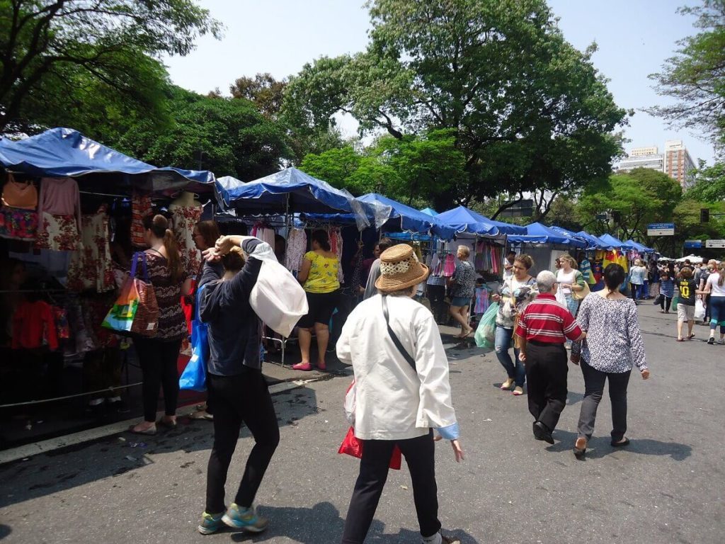 Foto que ilustra matéria sobre onde comprar roupas baratas em Belo Horizonte mostra a Feira Hippie da cidade, com barracas com coberturas azuis repletas de roupas à venda e pessoas circulando ao lado delas.