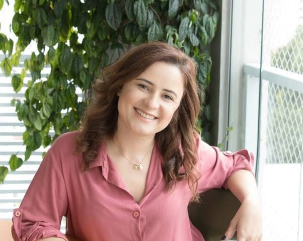Uma mulher de cabelos castanhos ondulados, vestindo uma blusa rosa, está sentada sorrindo em uma sala iluminada com plantas verdes frondosas e luz natural, acentuada por sutis elementos de decoração gótica ao fundo.
