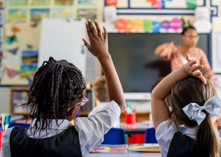 Uma menina, em sala de aula, levanta a mão para tirar dúvidas com a professora.