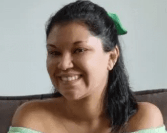 Uma mulher com longos cabelos escuros, usando um laço verde e uma blusa verde-claro tomara que caia, está sorrindo enquanto está sentada em um sofá marrom em uma sala bem iluminada.
