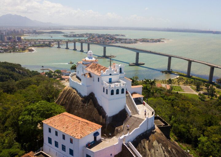 Foto mostra o Convento da Penha visto de cima, no alto de um morro, com a baía e a cidade de Vila Velha ao fundo.