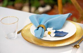 Imagem de uma mesa posta com sousplat dourado, guardanapo de pano na cor azul, flor decorativa e copo de vidro.