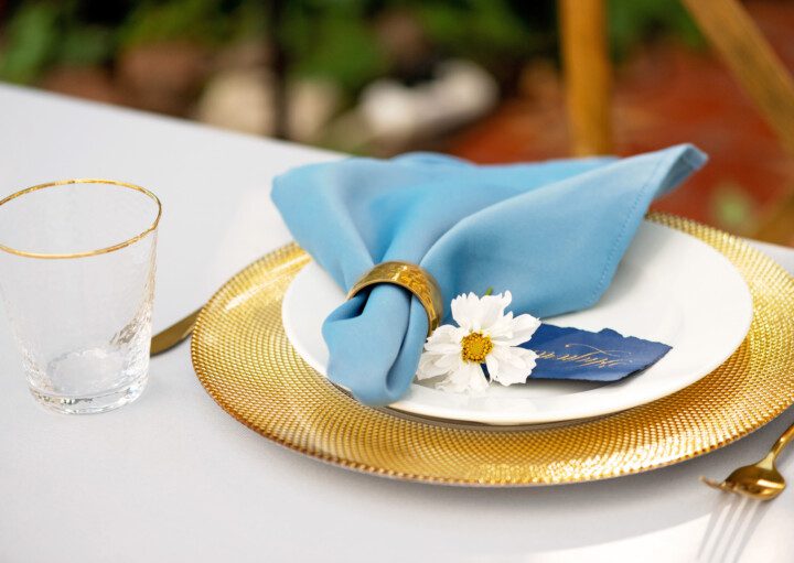 Imagem de uma mesa posta com sousplat dourado, guardanapo de pano na cor azul, flor decorativa e copo de vidro.