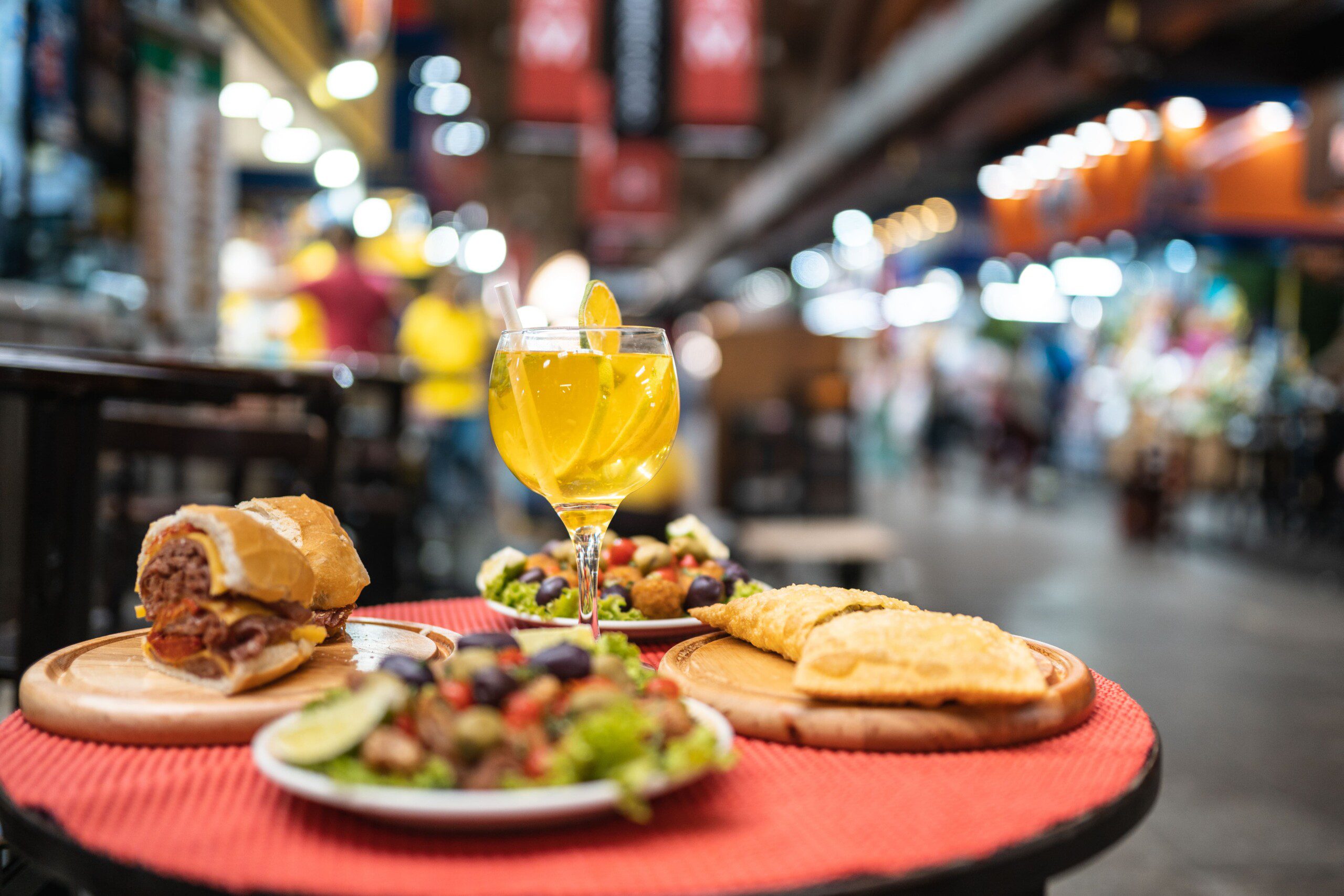 Imagem de lanches tradicionais no Mercadão de SP como sanduíche de mortadela, frutas cristalizadas e uma taça de drink. 