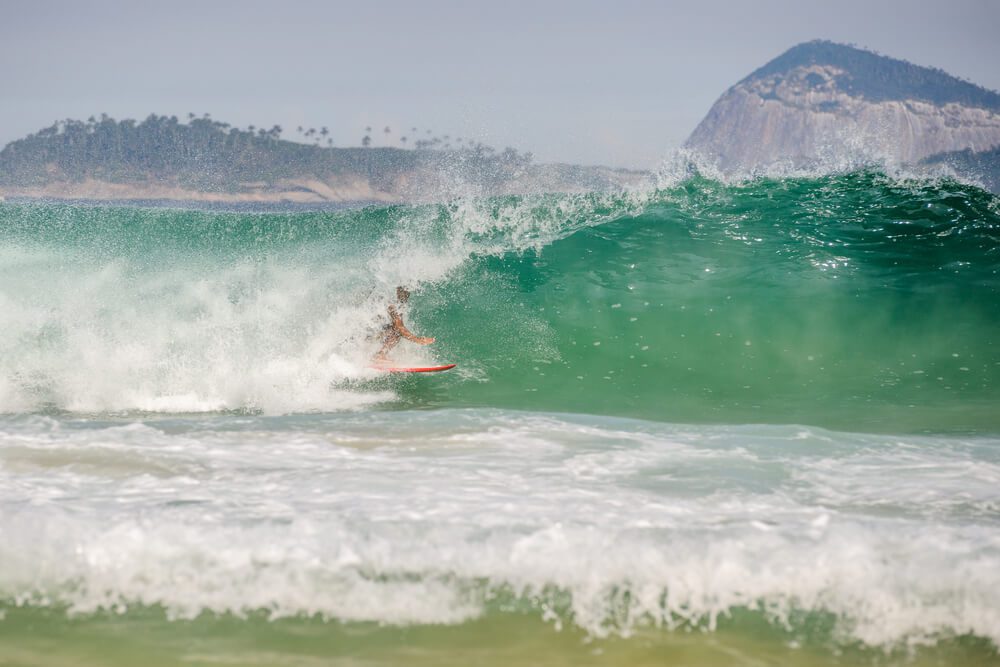 Foto que ilustra matéria sobre ilha no Rio de Janeiro mostra um surfista em ação pegando uma onda em primeiro plano. E ao fundo aparece uma grande rocha que faz parte das Ilhas Cagarras.