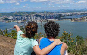 Foto que ilustra matéria sobre o Morro do Moreno mostra um casal sentado de costas olhando a vista da Baía de Vitória