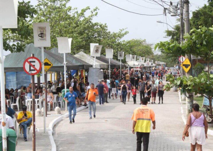 Foto que ilustra matéria sobre a Praia das Pedrinhas mostra a rua adjascente repleta espaços cobertos por toldos onde muitas pessoas circulam ou estão sentadas