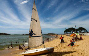 Foto que ilustra matéria sobre Praias de Vila Velha mostra a faixa de areia da Praia da Sereia, com destaque, em primeiro plano, para um pequeno barco a vela parado na areia repleta de cadeiras e barracas. Parte do mar, com alguns banhistas dentro, aparecem do lado esquero da tela.