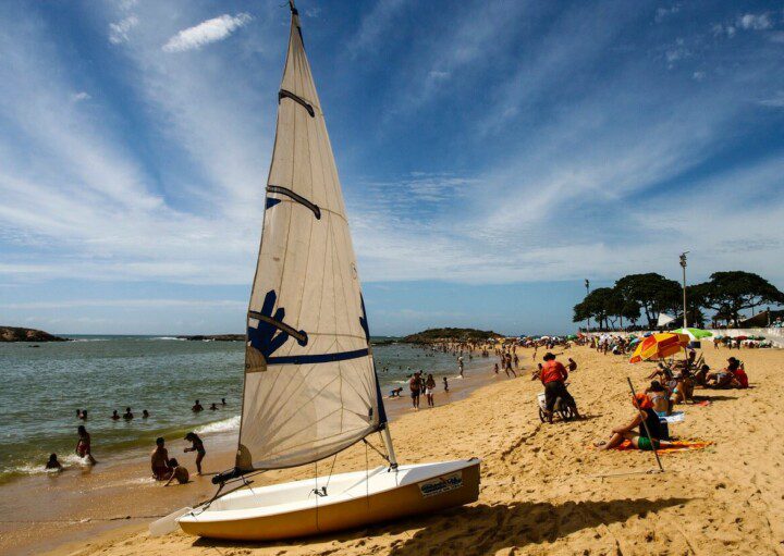 Foto que ilustra matéria sobre Praias de Vila Velha mostra a faixa de areia da Praia da Sereia, com destaque, em primeiro plano, para um pequeno barco a vela parado na areia repleta de cadeiras e barracas. Parte do mar, com alguns banhistas dentro, aparecem do lado esquero da tela.