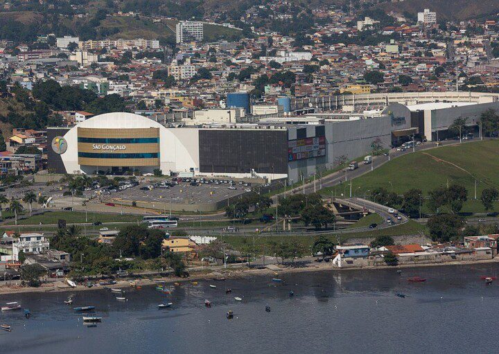 Foto mostra uma visão do alto e de longe do São Gonçalo Shopping, com uma parte da Baía de Guanabara aparecendo em primeiro plano, o shopping no Centro e a cidade atrás.