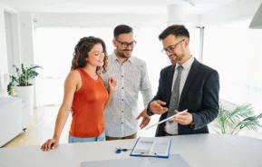 Imagem de um casal formado por um homem e uma mulher em um imóvel olhando uns papéis apresentados por um corretor de imóveis para ilustrar matéria sobre carta de crédito