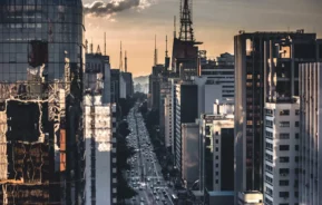 Imagem da vista de um mirante na Avenida Paulista mostra pôr do sol na avenida movimentada ladeada por edifícios altos e modernos, alguns com fachadas de vidro que refletem a luz. Várias antenas e torres erguem-se acima da linha do horizonte sob um céu parcialmente nublado