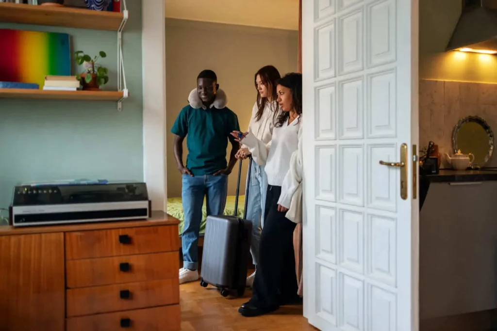 Imagem de três pessoas com uma mala estão na porta de seu quarto de aluguel temporada. Uma pessoa usa um travesseiro de viagem. O espaço aconchegante tem uma cômoda, um toca-discos e prateleiras com livros e uma planta.