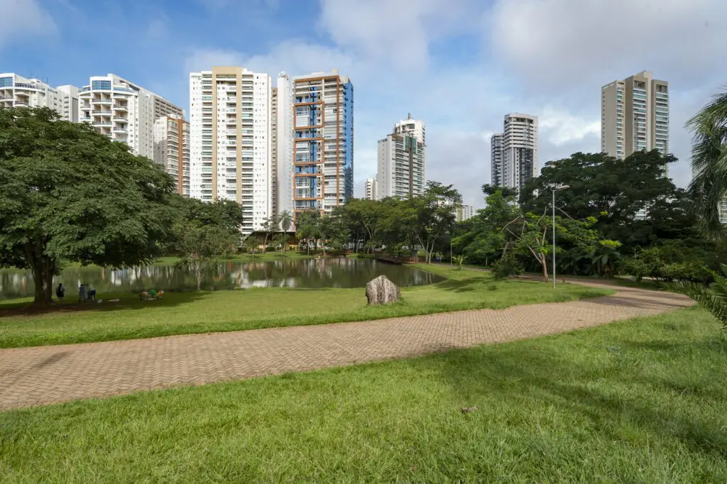 Um parque da cidade de Goiânia com uma pista de caminhada, grama verde, árvores grandes, um lago e prédios de apartamentos altos e modernos ao fundo, sob um céu parcialmente nublado - um local ideal quando se busca o que fazer em Goiânia.