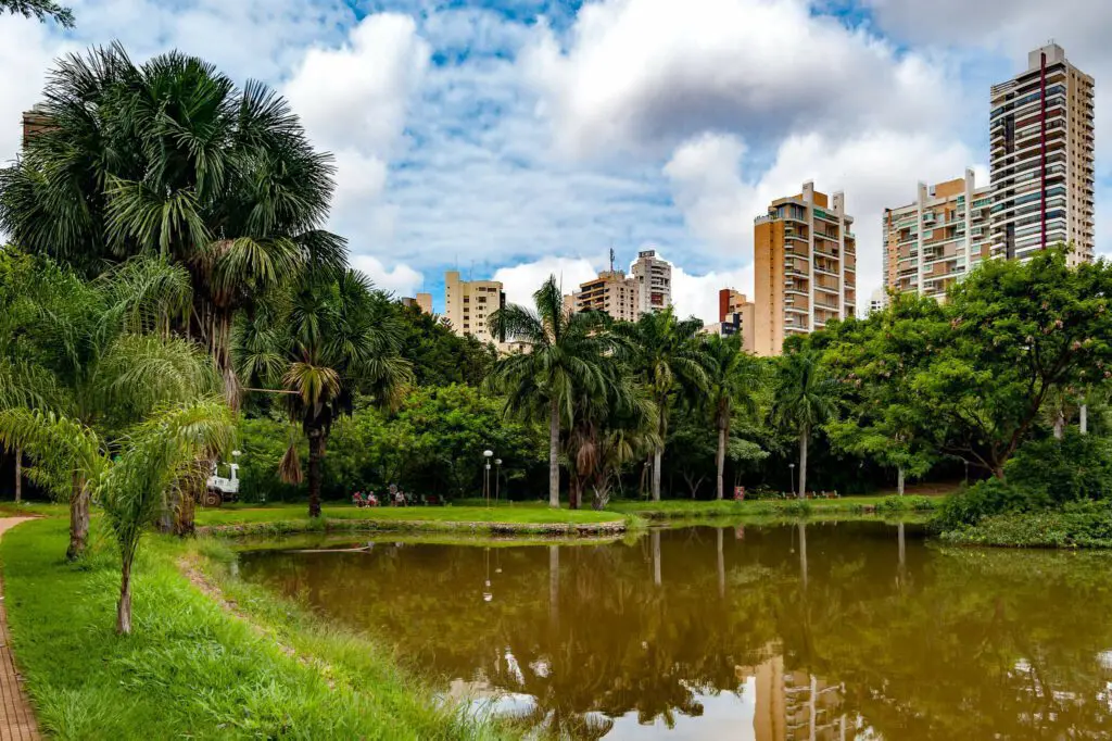 Um pequeno lago cercado por árvores verdes e palmeiras em um parque da cidade, com altos prédios de apartamentos ao fundo sob um céu parcialmente nublado - um local convidativo para quem está explorando o que fazer em Goiânia.