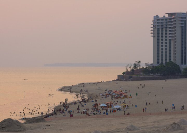 Foto da Praia de Ponta Negra durante o pôr do sol. Há pessoas na areia e prédios ao fundo.