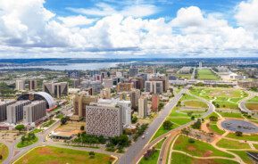 Foto que ilustra matéria sobre o Eixo Monumental mostra a região do alto, com alguns prédios à esquerda, uma avenida no meio e um céu azul com nuvens ao fundo