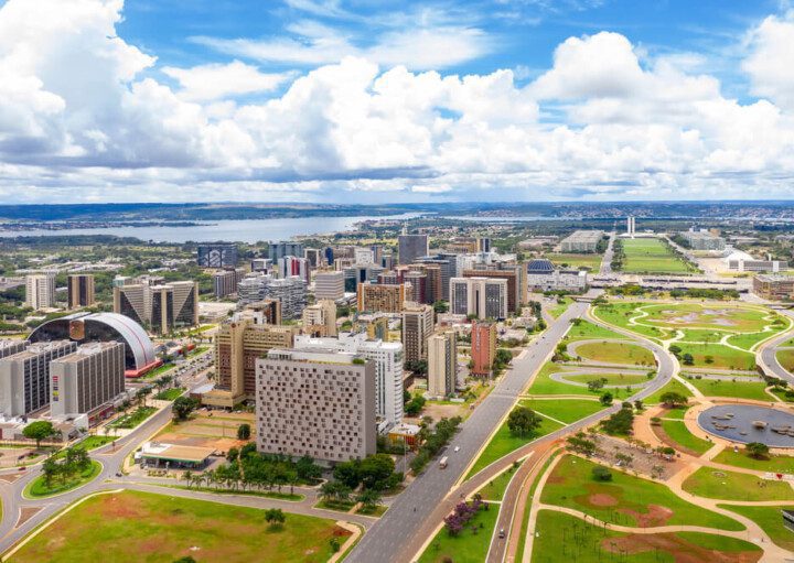 Foto que ilustra matéria sobre o Eixo Monumental mostra a região do alto, com alguns prédios à esquerda, uma avenida no meio e um céu azul com nuvens ao fundo