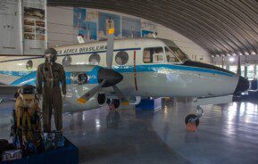 Foto que ilustra matéria sobre o Museu Aeroespacial Brasileiro mostra o interior de um galpão com um pequeno avião. Ao lado dele se encontra um manequim com roupas de aviação e outros equipamentos.