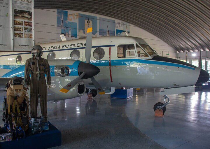 Foto que ilustra matéria sobre o Museu Aeroespacial Brasileiro mostra o interior de um galpão com um pequeno avião. Ao lado dele se encontra um manequim com roupas de aviação e outros equipamentos.