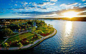 Foto que ilustra matéria sobre o Lago Paranoá mostra o espelho d'água ao Centro, com um pôr do sol à direita e uma das margens do lago, com uma área verde, à esquerda.