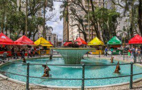 Foto que ilustra matéria sobre a Praça Osório, em Curitiba, mostra o chafariz localizado no centro da praça cercado de barraquinhas de comida