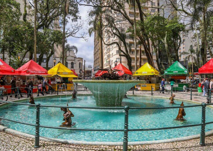 Foto que ilustra matéria sobre a Praça Osório, em Curitiba, mostra o chafariz localizado no centro da praça cercado de barraquinhas de comida