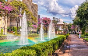 Foto que ilustra matéria sobre a Praça da Liberdade em Belo Horizonte mostra um trecho da praça onde há uma fonte de água com chafarizes, um coreto e o Edifício JK ao fundo.