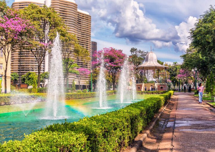 Foto que ilustra matéria sobre a Praça da Liberdade em Belo Horizonte mostra um trecho da praça onde há uma fonte de água com chafarizes, um coreto e o Edifício JK ao fundo.