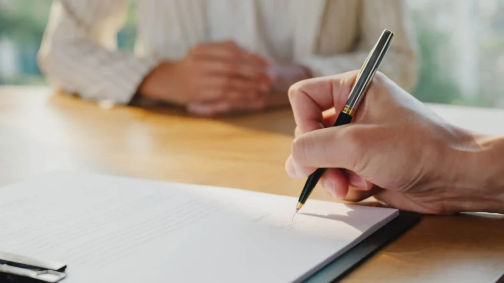 Imagem de uma mão segurando uma caneta, assinando uma renovação de contrato de aluguel em uma prancheta, com outra pessoa sentada do outro lado da mesa ao fundo, com as mãos cruzadas