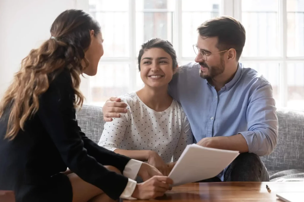 Imagem de um casal sorridente sentado em um sofá, analisando documentos sobre renovação de contrato de aluguel com uma mulher. O homem tem o braço em volta da mulher e todos parecem felizes, sugerindo uma reunião bem-sucedida em uma sala iluminada e moderna