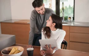 Imagem de uma mulher sentada em uma mesa de cozinha segurando um tablet olha para cima e sorri para um homem ao seu lado para ilustrar matéria sobre a diferença entre escritura e registro