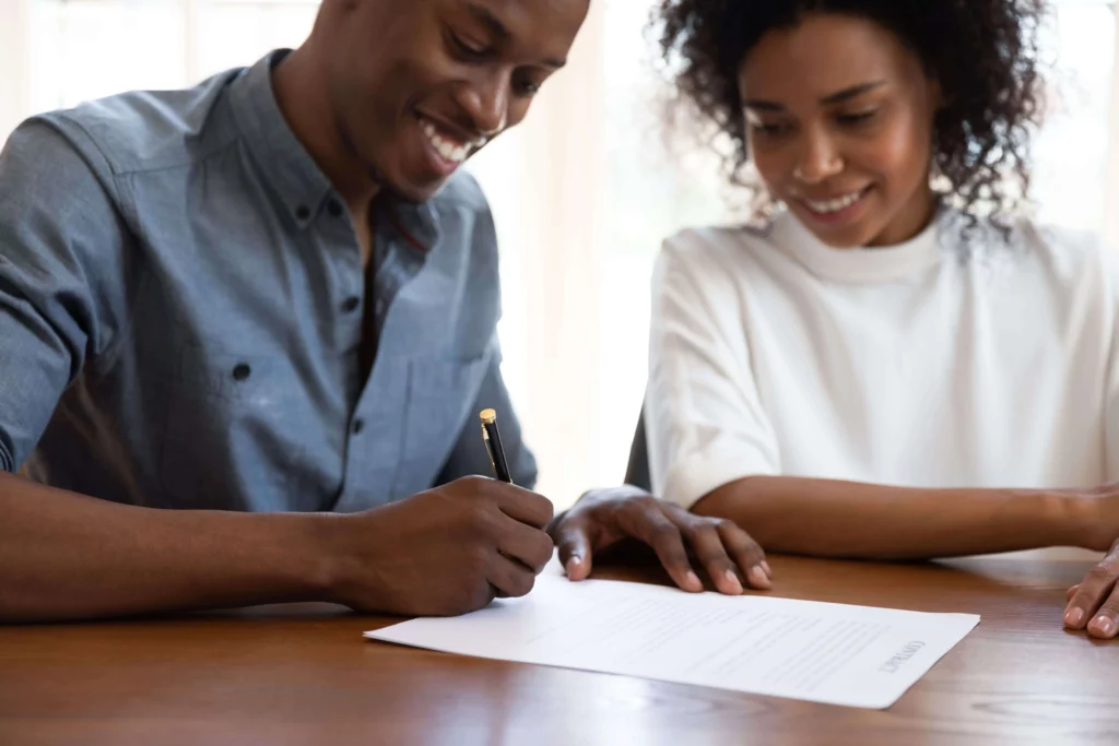 Imagem de um homem e uma mulher sorridentes sentados à mesa; o homem está assinando um documento com uma caneta enquanto a mulher observa, ambos parecendo felizes e engajados durante uma renovação de contrato de aluguel