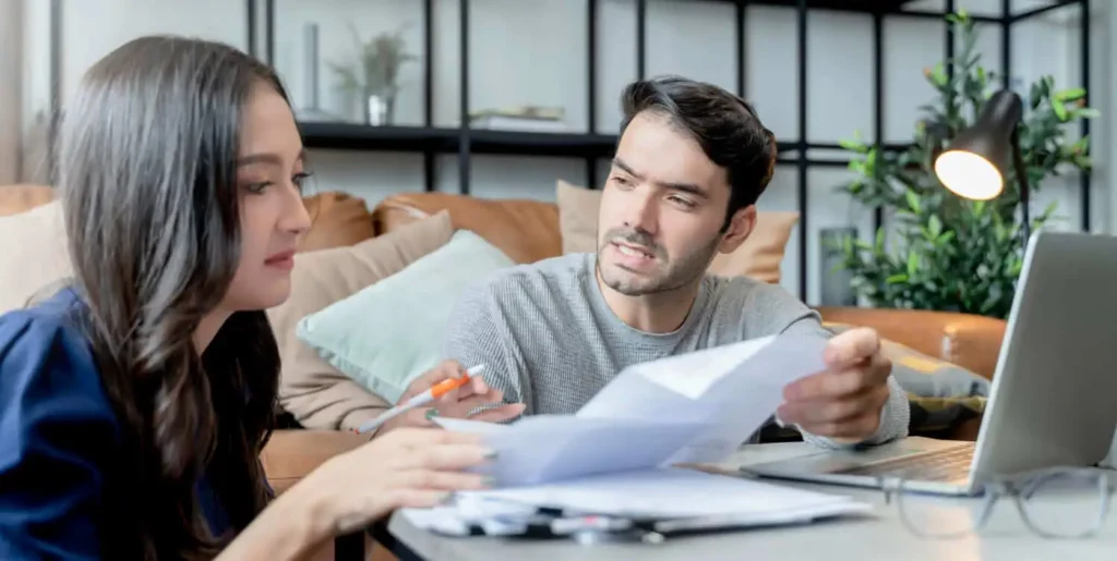 Imagem de um homem e uma mulher sentados à mesa com papéis e um laptop, em sua aconchegante e moderna sala de estar, para ilustrar matéria sobre qual a diferença entre escritura e registro de imóveis.