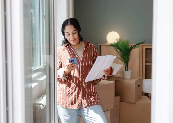 Imagem de uma mulher em um ambiente interno de uma casa segurando papéis e olhando para seu telefone, sorrindo, cercada por caixas de papelão e um vaso de planta
