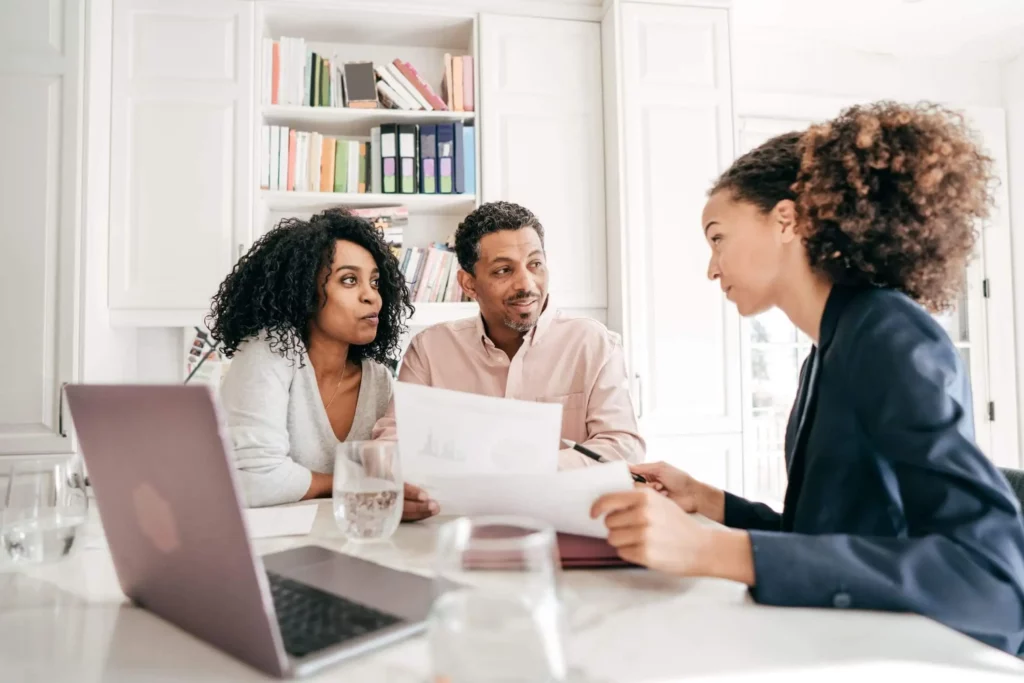 Imagem de três pessoas sentamdas à mesa em um escritório bem iluminado. Uma mulher de blazer segura papéis e conversa com um homem e uma mulher à sua frente. Um laptop, copos de água e estantes de livros estão ao fundo
