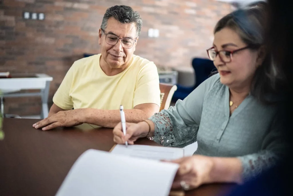 Imagem de um homem mais velho, de camisa amarela, sentado em uma mesa observando uma mulher de óculos e blusa azul assinar um documento de renovação de contrato de aluguel. O cenário parece ser um ambiente interno, com uma parede de tijolos ao fundo