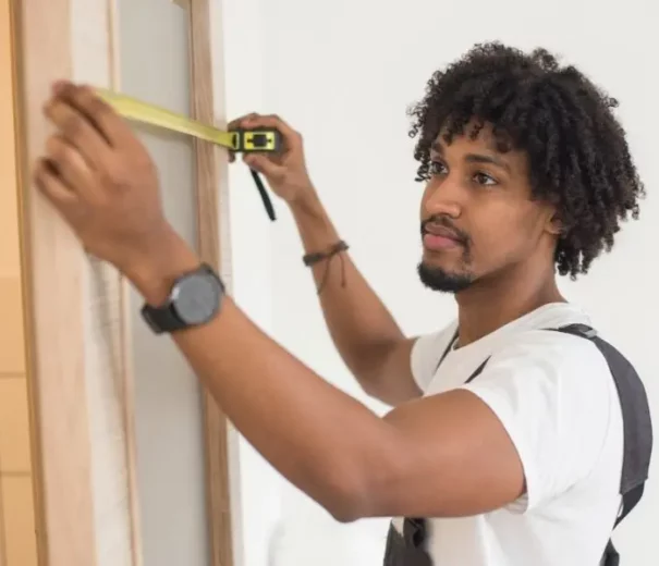 Foto que ilustra matéria sobre como calcular o metro quadrado mostra um homem jovem abrindo uma trena em uma parede.