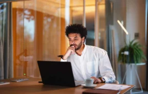 Imagem de um homem de cabelos cacheados e barba, vestido com uma camisa branca, sentado em uma mesa, olhando atentamente para a tela de um laptop enquanto pesquisa sobre due diligence imobiliária. Uma xícara, papéis e caneta estão sobre a mesa; a sala é moderna e bem iluminada.