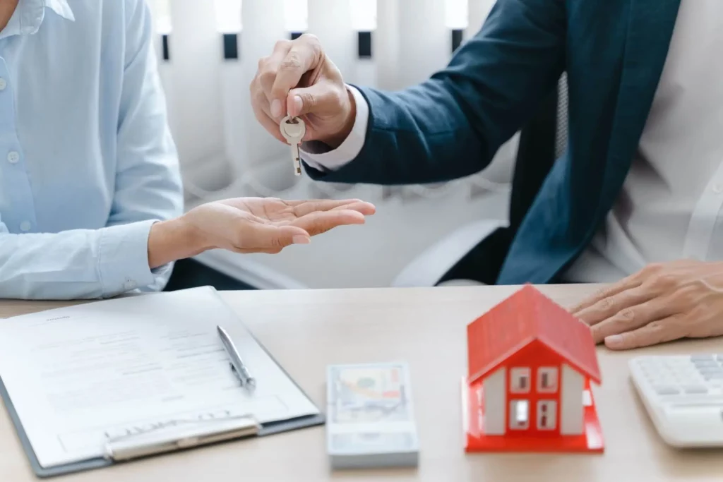 Imagem de uma pessoa entregando a chave de uma casa para outra em uma mesa com um contrato, uma pilha de dinheiro e um modelo de casa vermelha em foco em cima de uma mesa