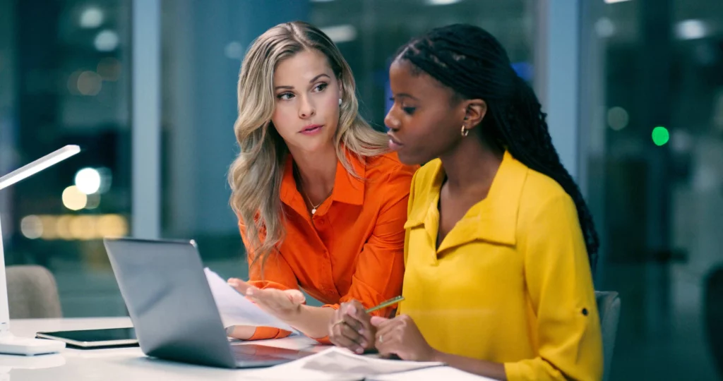 Imagem de duas mulheres sentadas em uma mesa trabalhando juntas. Uma, de camisa laranja, está explicando a due diligence imobiliária enquanto segura papéis; a outra, de camisa amarela, faz anotações em um bloco de notas ao lado de um laptop.