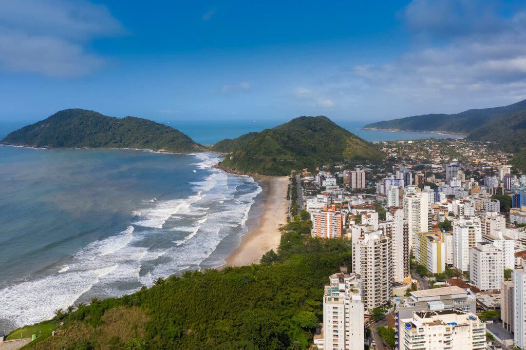 Imagem aérea da praia. É possível ver o céu e mar azuis, um pouco de mata atlântica nas pontas da faixa de areia e prédios ao redor.