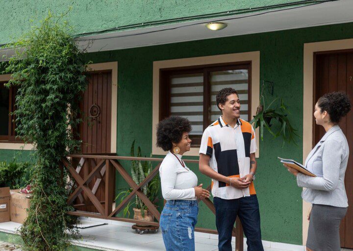 Foto que ilustra matéria sobre casa de vila mostra um casal sorridente conversando com uma corretora de imóveis em frente a uma casa verde