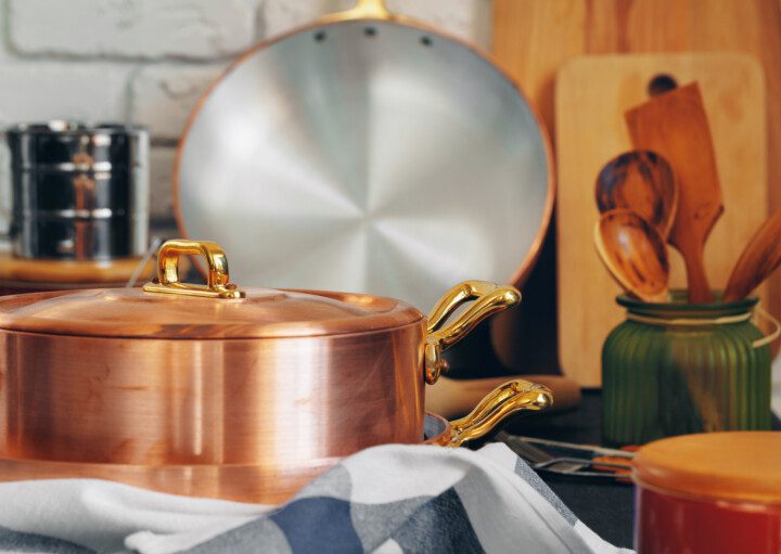 Imagem de uma panela de cobre com utensílios de madeira perto, como talheres e tábuas, em cima de uma mesa estampada com uma toalha quadriculada